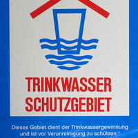 Schild - Trinkwasserschutzgebiet - blauer Hintergrund, rote Schrift - symbolisch: überdachte Tasse steht halb im Wasser
